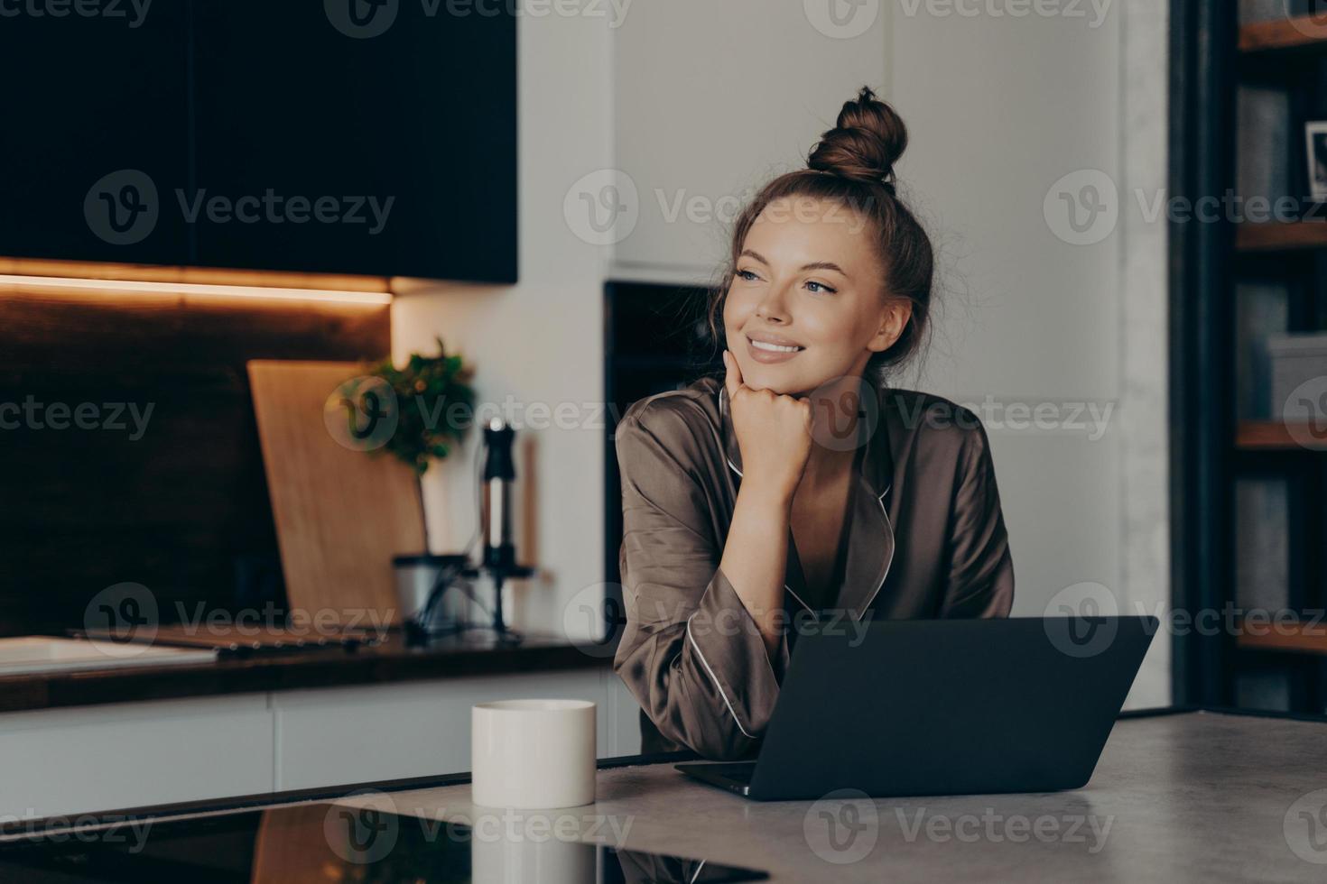 joven mujer independiente feliz en pijamas acogedores trabajando desde casa en una computadora portátil foto