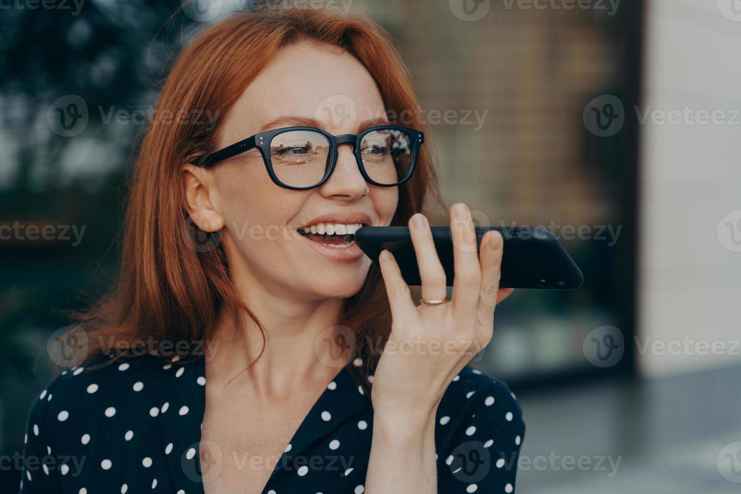 una mujer de negocios pelirroja sonriente que usa un asistente de voz digital virtual mientras está de pie al aire libre foto