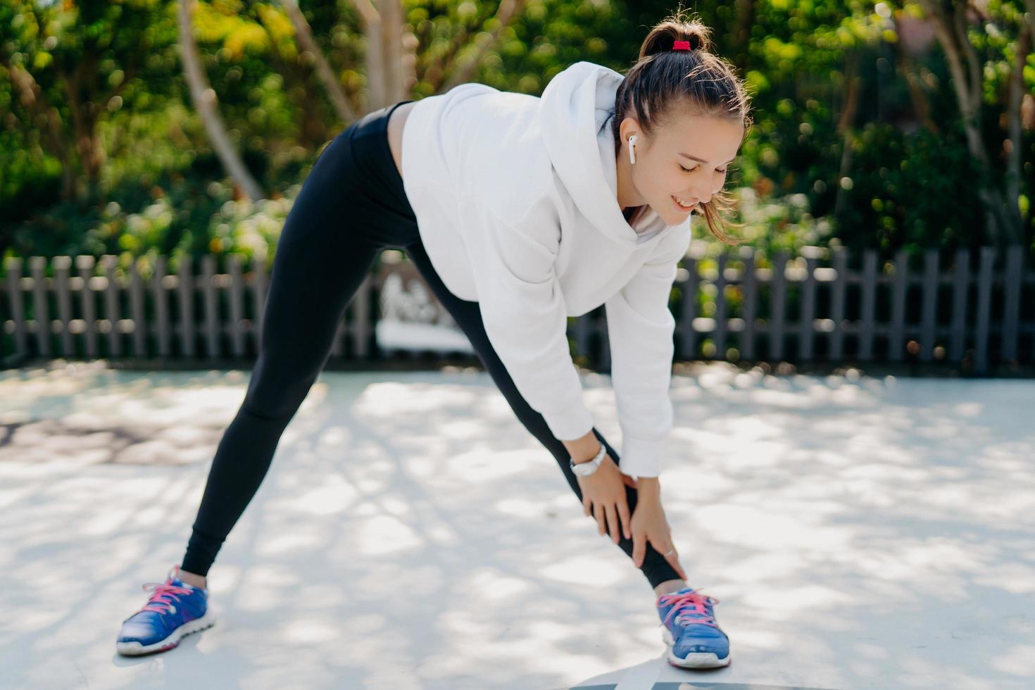 mujer adulta joven motivada practica deporte con regularidad se estira de la mano a los pies se inclina hacia abajo tiene los pies separados al ancho de los hombros usa ropa deportiva disfruta entrenar actividades deportivas al aire libre para perder peso foto