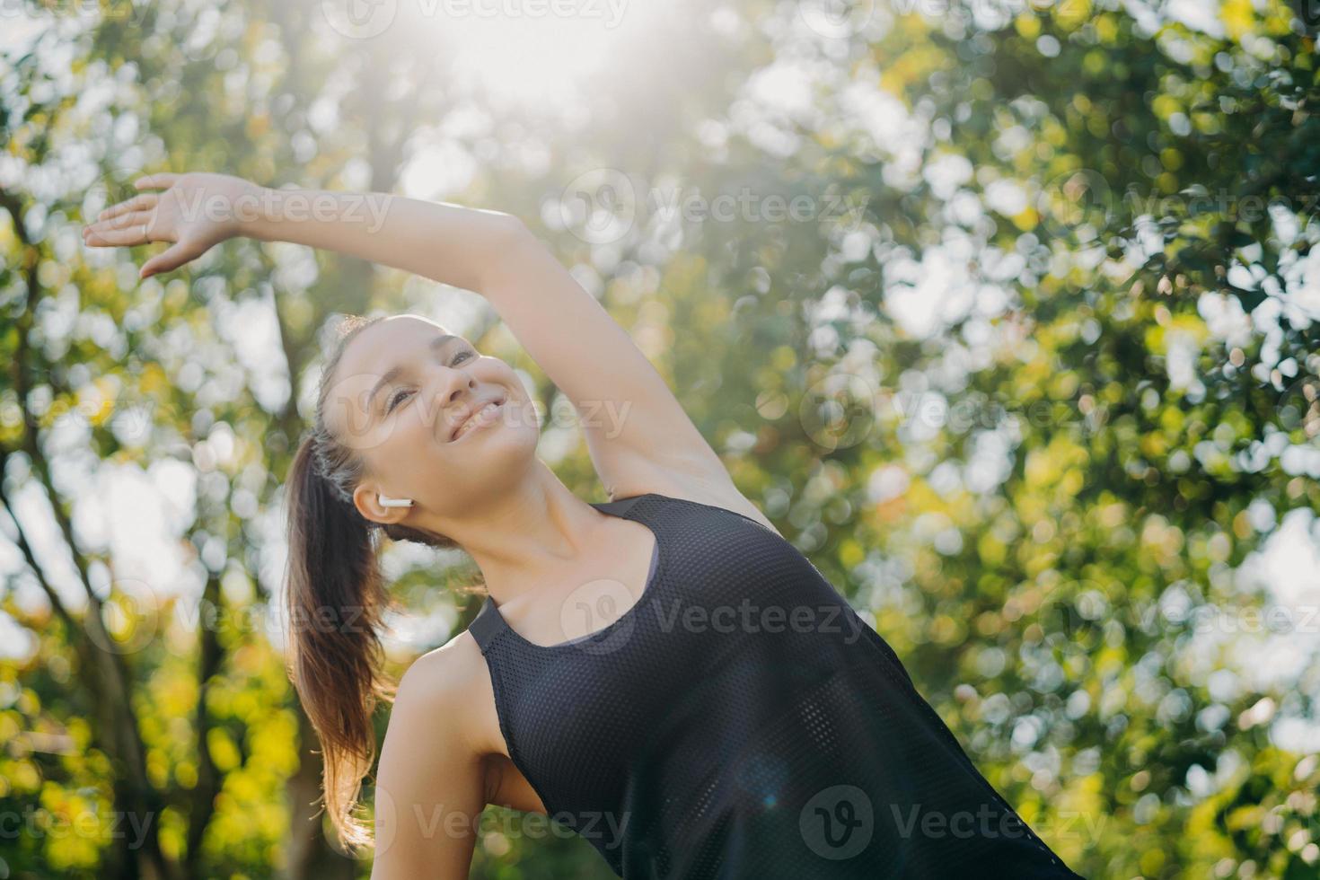 Cheerful sporty woman with pony tail leans aside exercises outdoor against trees enjoys sunny day listens music via wireless earphones has morning training seession performs warm up cardio workout photo
