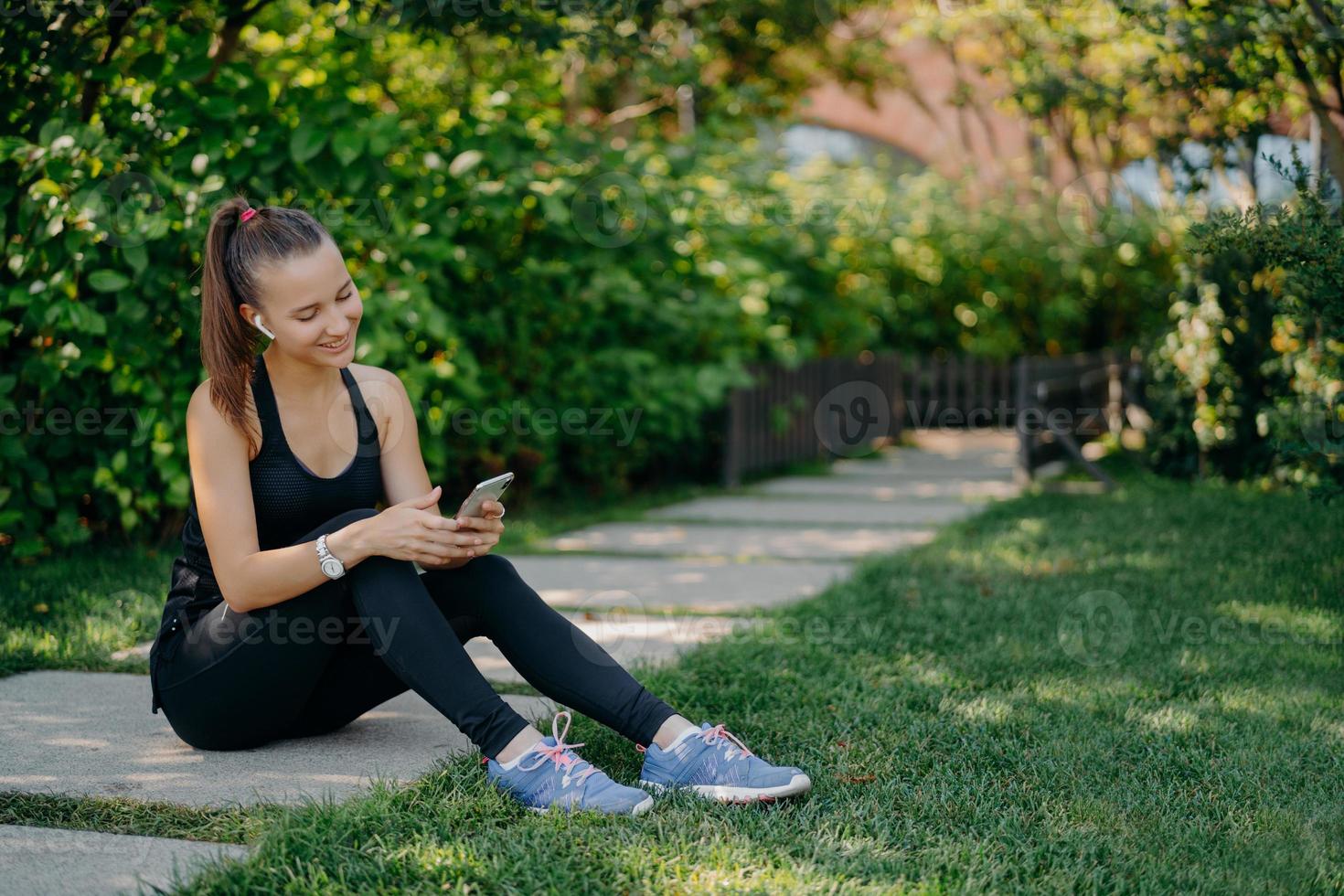 Pleased athletic woman in sportsclothes smiles cheerfully surfs mobile phone while listens music via wireless earphones takes break after sunny morning workout uses app for activity tracking photo