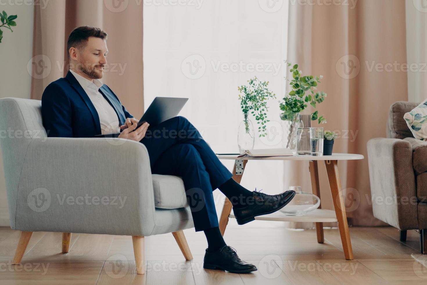 Successful businessman in formal wear sits in comfortable armchair uses modern laptop prepares online project poses against home interior analyzes financial indicators controls company development photo