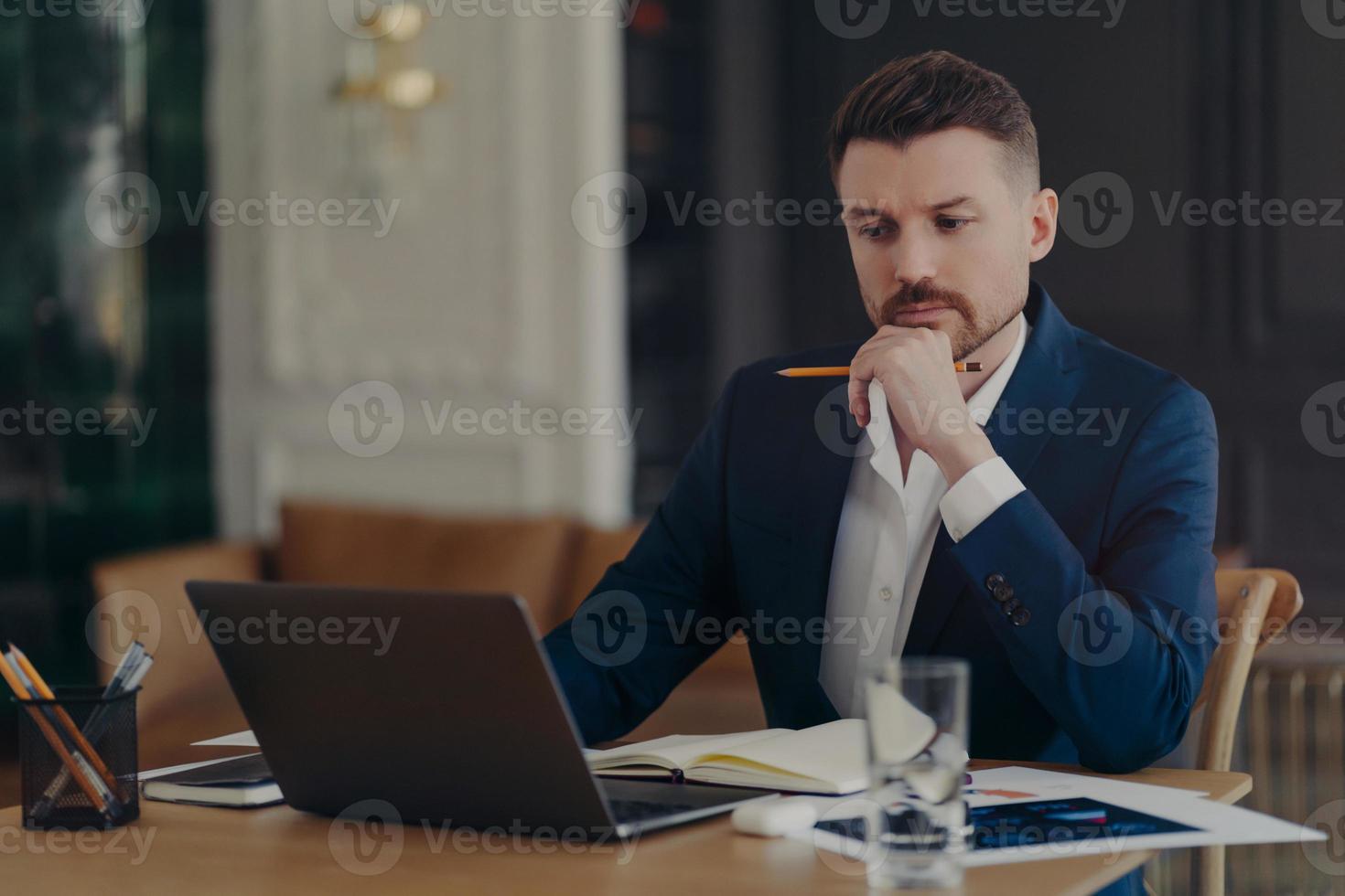 Serious male entrepreneur holds chin makes notes in diary works on laptop computer makes money online dressed formally writes information. Confident managing director involved in working process photo