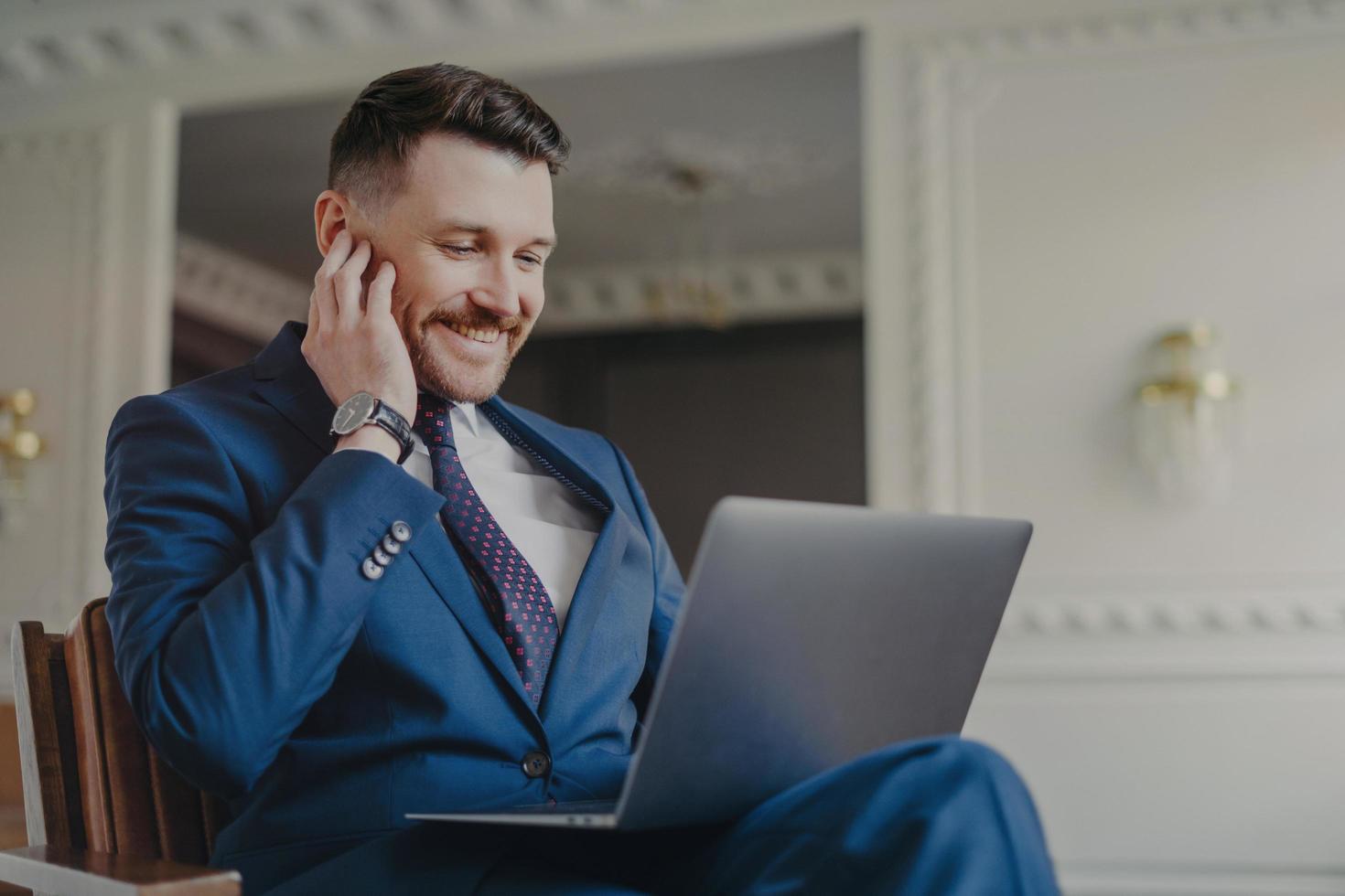 Cheerful corporate employer uses laptop computer reads news online or financial publication connected to 4g internet sits in comfortable armchair dressed in blue suit arranges formal meeting photo