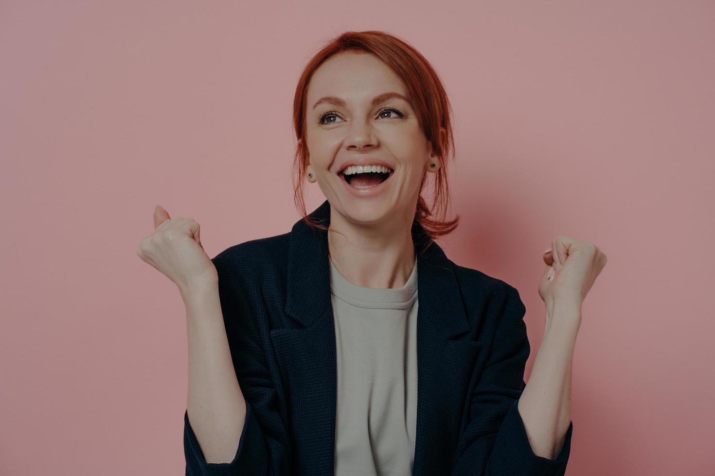 retrato de mujer pelirroja feliz y emocionada con los puños levantados celebrando la victoria, aislada en rosa foto