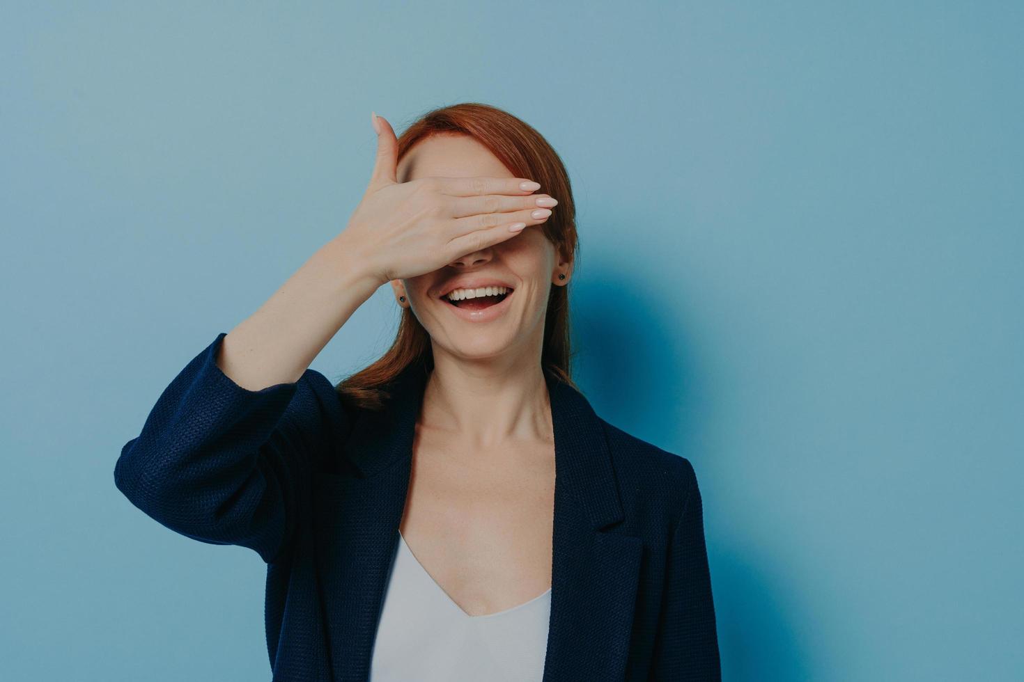 Playful funny ginger girl covering her eyes with palm isolated on blue background, isolated on blue photo