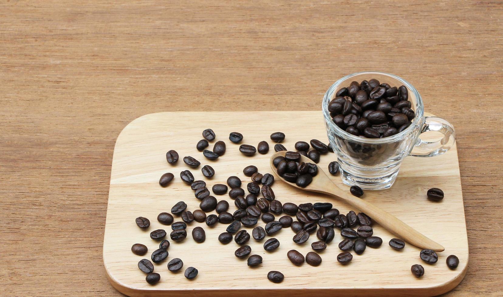 granos de café tostados marrones en una taza de espresso sobre una tabla de madera con una cuchara de madera y un montón de granos de café sobre una mesa rústica de madera foto