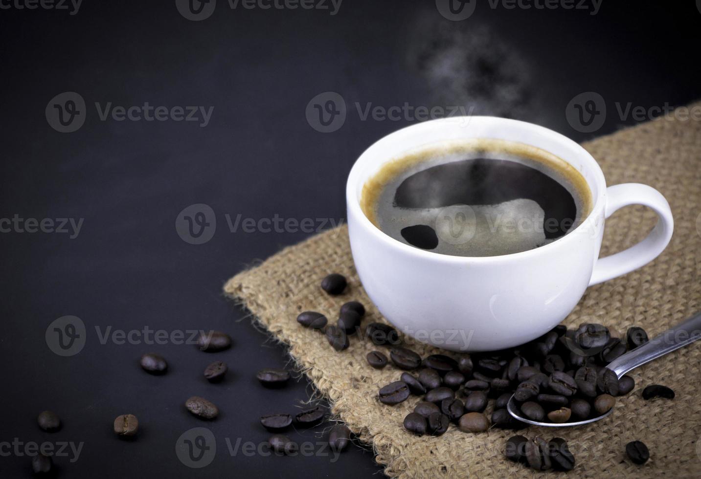 High angle view of a white ceramic cup of black hot Americano coffee with steam on a sackcloth with a pile of roasted coffee beans on black background. photo