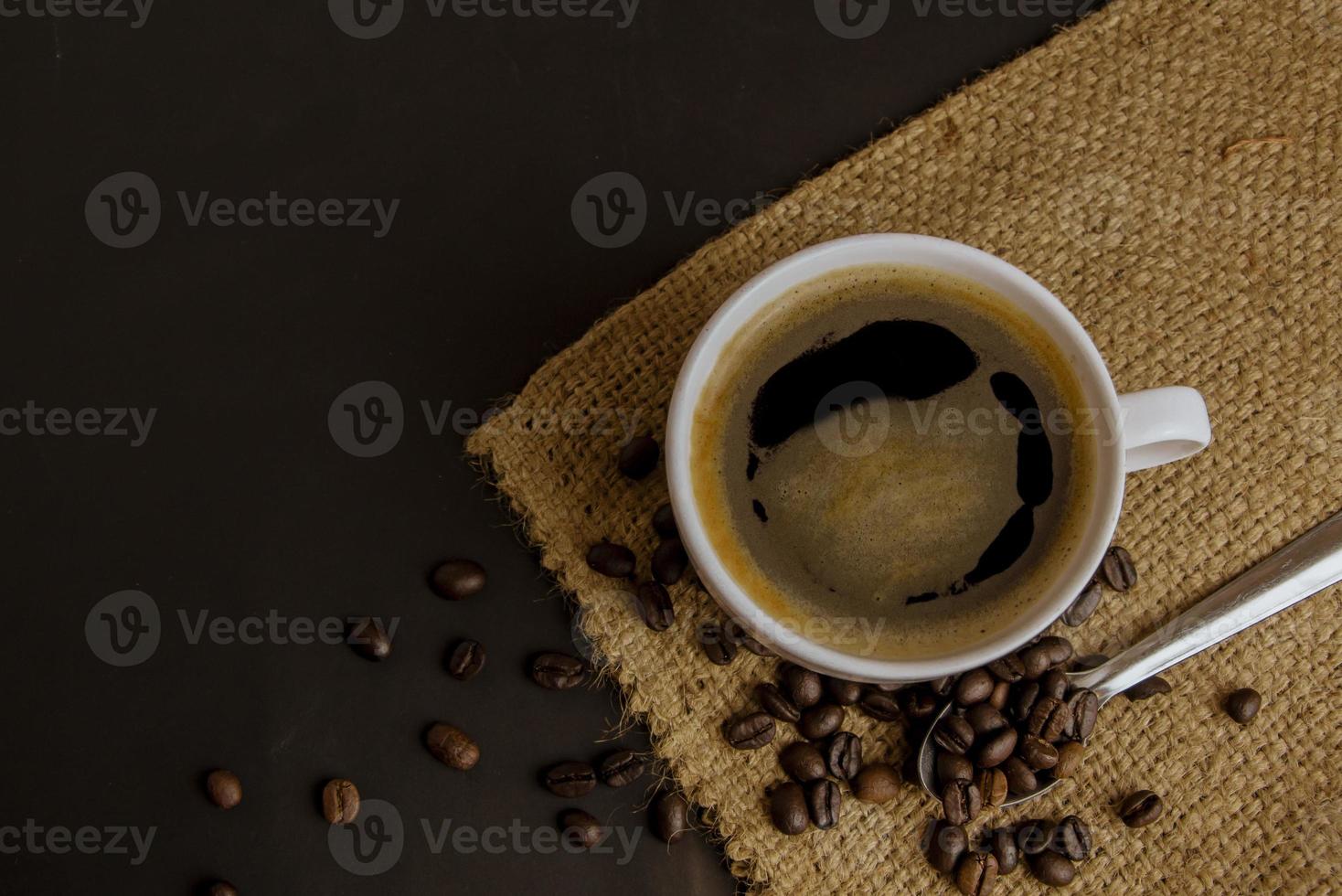 Top view of a white ceramic cup of black Americano hot coffee on sackcloth and a dark background with a pile of roasted coffee beans photo