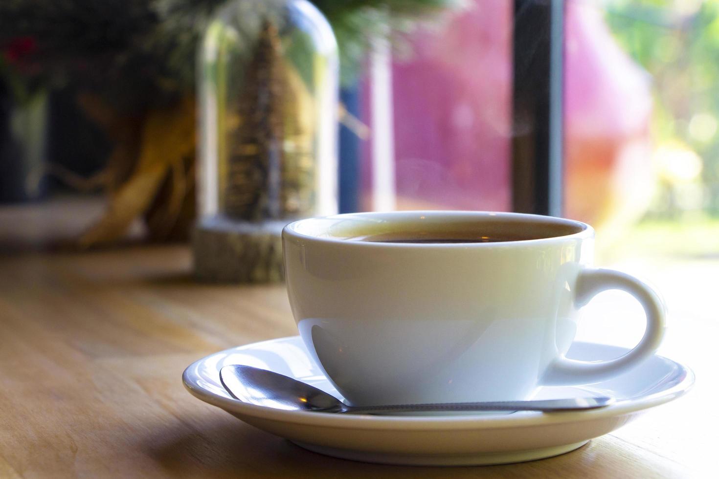 A white ceramic cup of hot black Americano coffee with light steam on top on a brown table in a coffee shop by the window. Drinks, relaxation, and holiday concept. photo