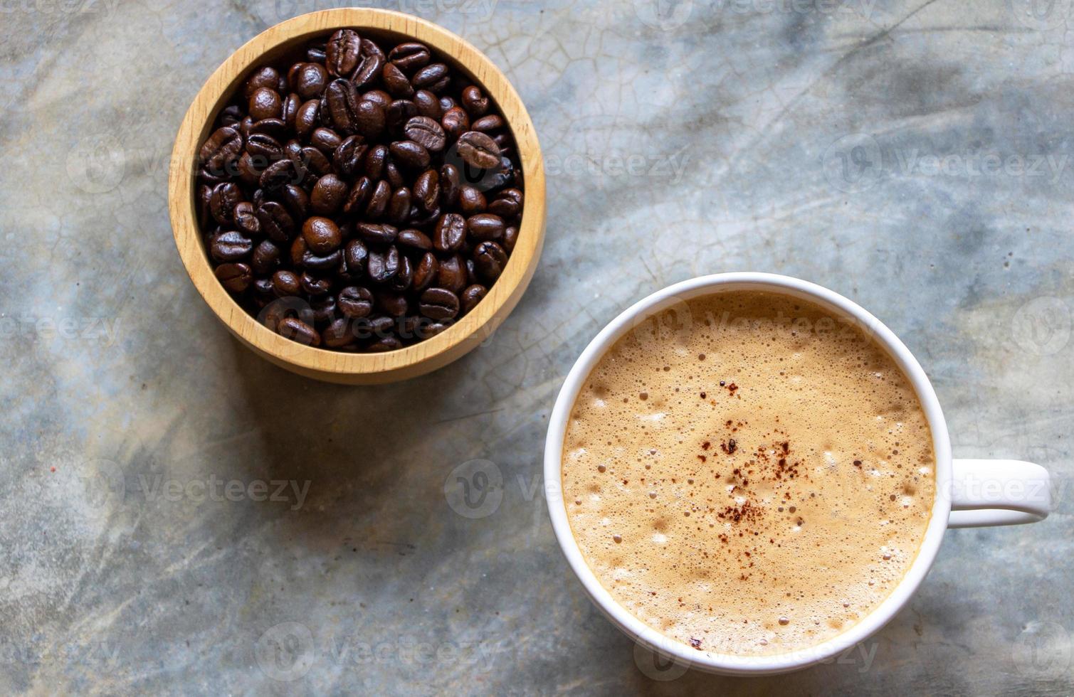 cup of mocha coffee with a nice foam on top on a concrete table and a wooden bowl of organic arabica coffee beans nearby. photo