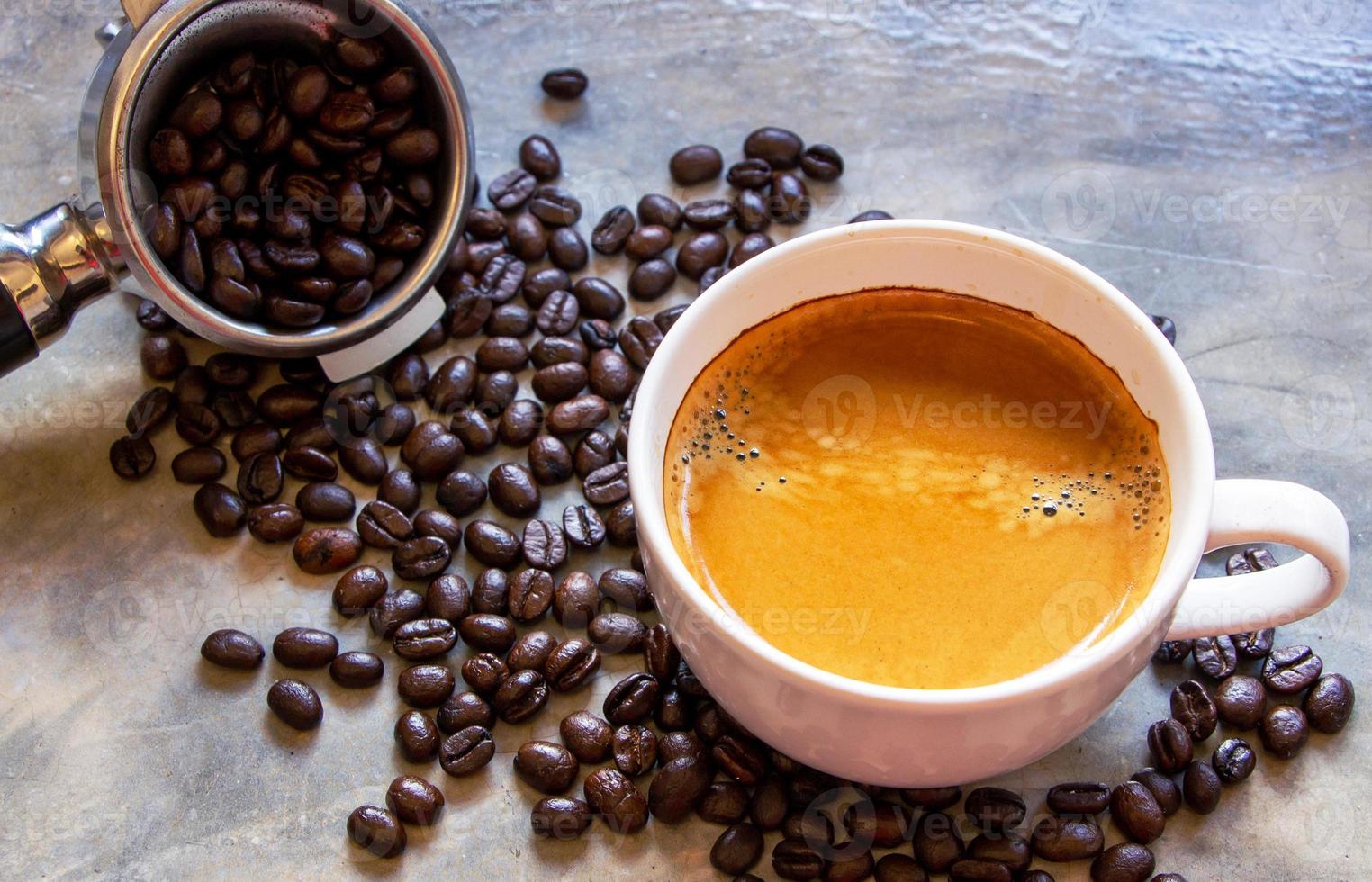 taza blanca de café americano con una bonita crema encima de una mesa de hormigón cerca de un portafiltros con granos de café arábica. centrarse en una taza de café foto