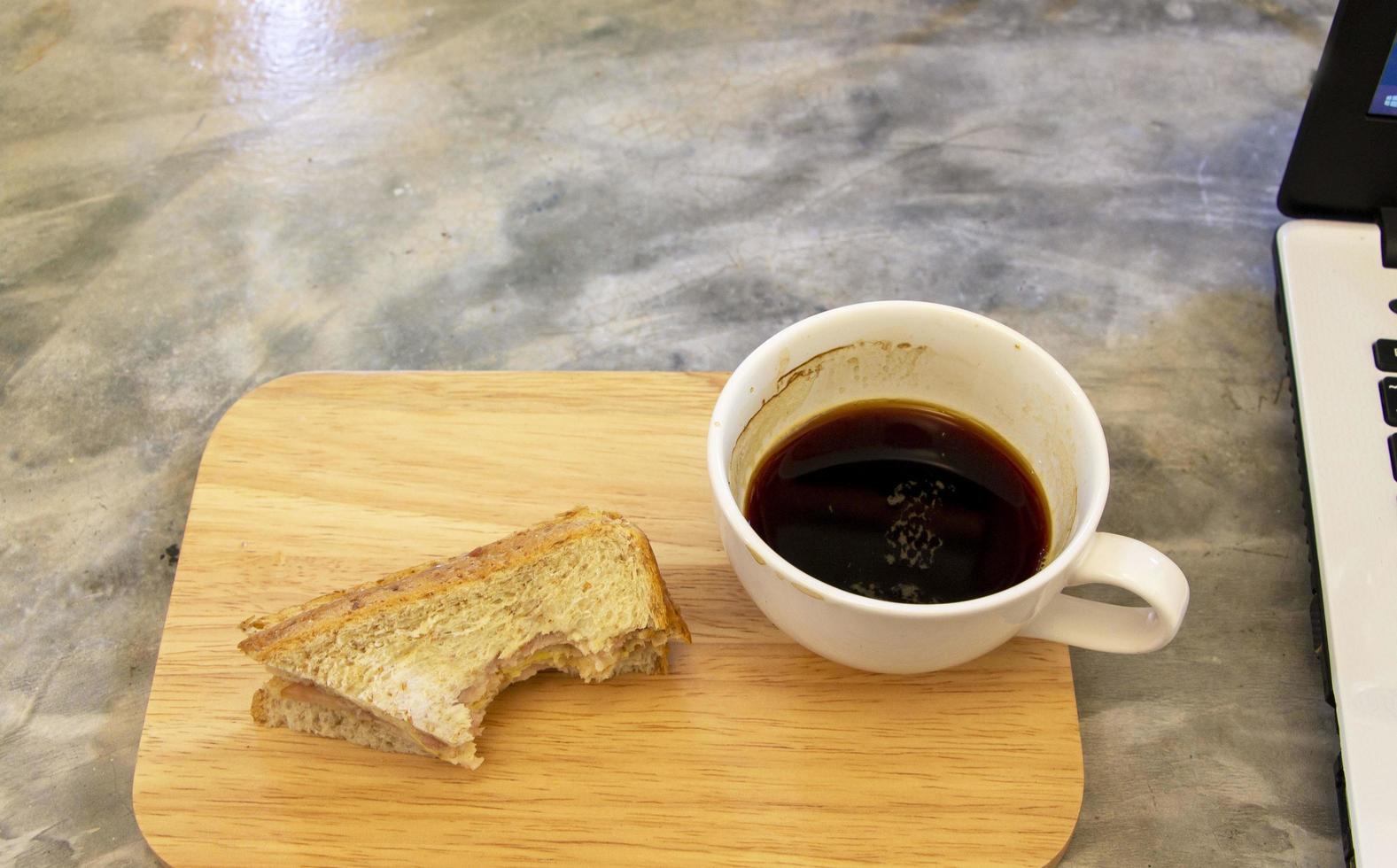 taza de café negro restante con un sándwich de queso de jamón mordido en una tabla de madera y mesa de hormigón gris foto