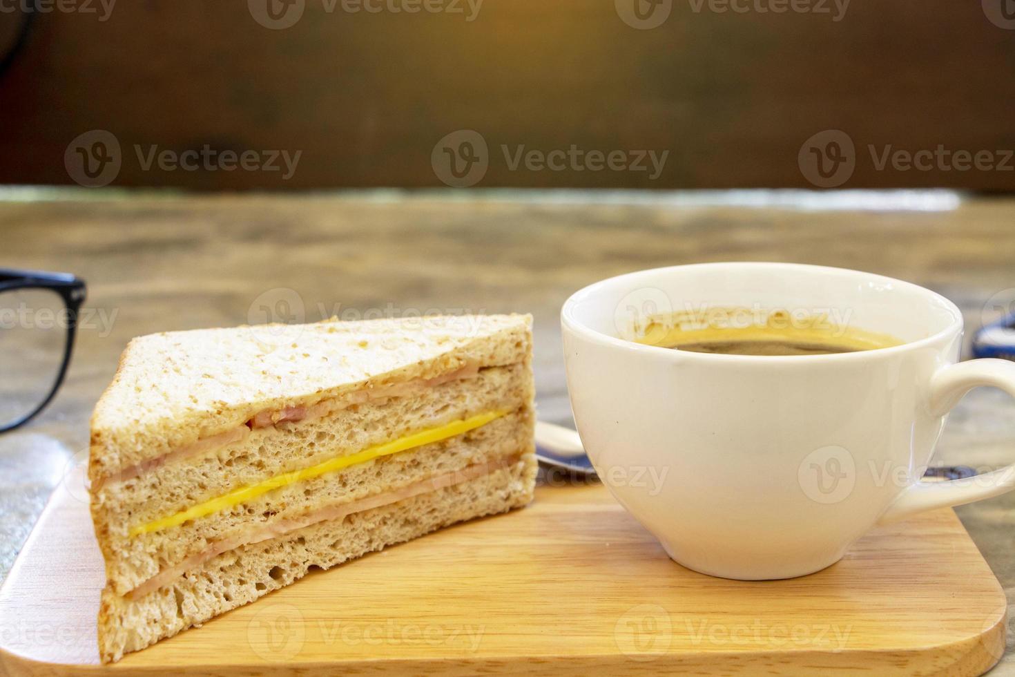 taza de café negro sobre una tabla de madera con sándwich de jamón y queso sobre una mesa rústica de hormigón foto