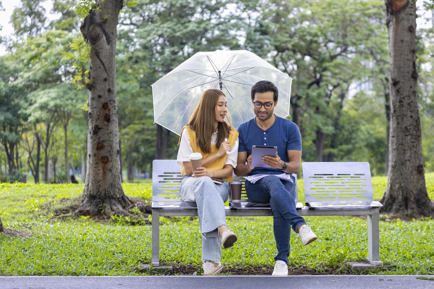 jóvenes asiáticos e indios disfrutan de relajarse bajo la lluvia juntos en el parque público mientras se sientan juntos en el banco durante el fin de semana foto