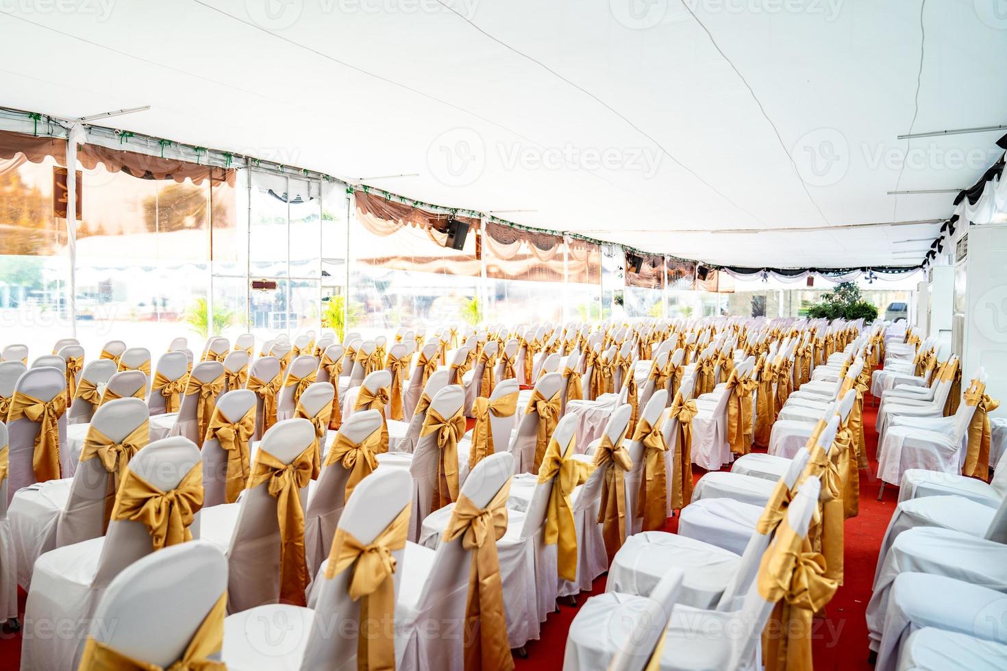 Temporary Air Conditioner indoor tent for outdoor event in the day. Inside Tents has red carpet and a lot of chair covered by white cloth and golde ribbon on it. photo