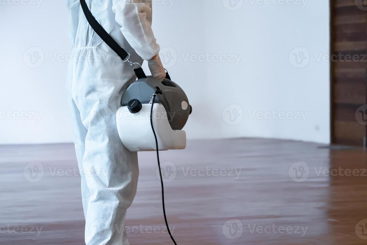 Professional technical man in prevention suit sprays sterilising solution by electrical spray machine on wood floor and white background with studio light. photo