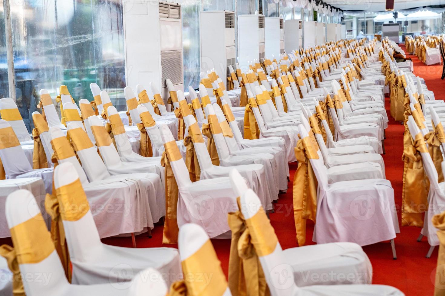 Temporary Air Conditioner indoor tent for outdoor event in the day. Inside Tents has red carpet and a lot of chair covered by white cloth and golde ribbon on it. photo