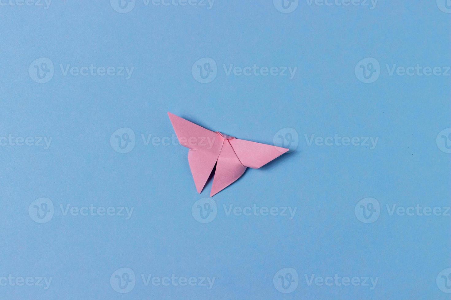 Pink origami butterfly folds out of paper. In center of a blue background. Education, pastime, hobbies, activities with children. Minimalistic background photo