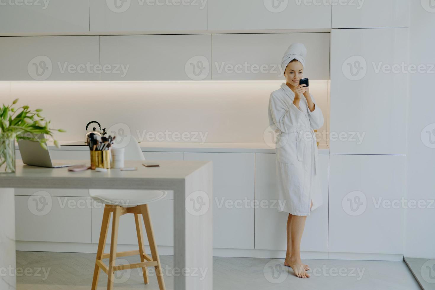 Full length shot of young beautiful woman dressed in white bathrobe applies face cream looks in mirror takes care of her skin poses near kitchen furniture stands bare feet. People beauty facial care photo
