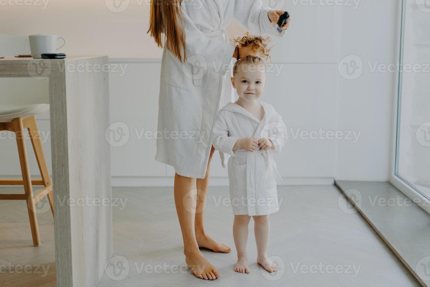 Faceless mother makes pony tail from daughters curly hair uses comb. Cute little three year old girl in white bathrobe after taking shower poses near mom at home. Children parenthood beauty time photo