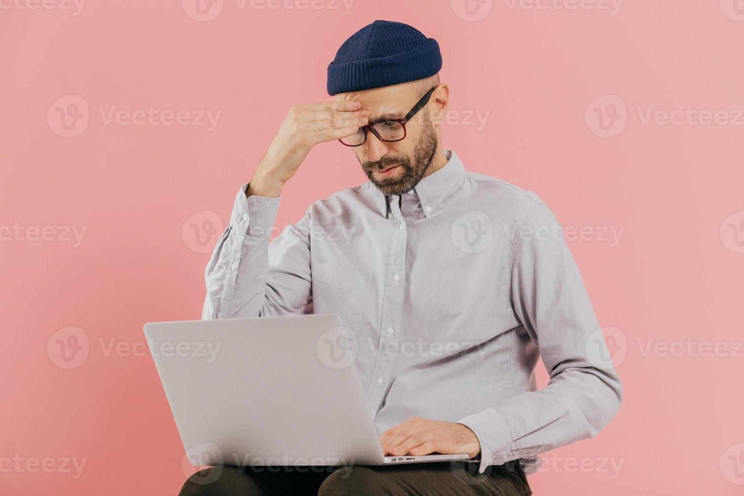 Male entrepreneur tries to make solution, keeps hand on head, tries to prepare financial report, focused in laptop computer, poses against pink background. Unshaven young man browses website photo
