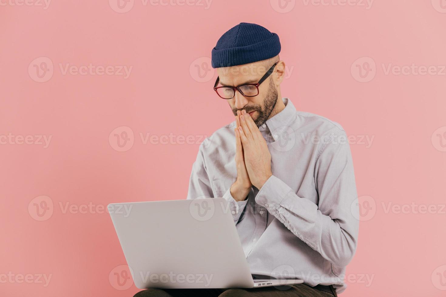 Professional male programmer keeps hans in praying gesture, looks at screen of laptop computer, believes in successful result of his work, wears formal shirt and hat, isolated over pink background photo