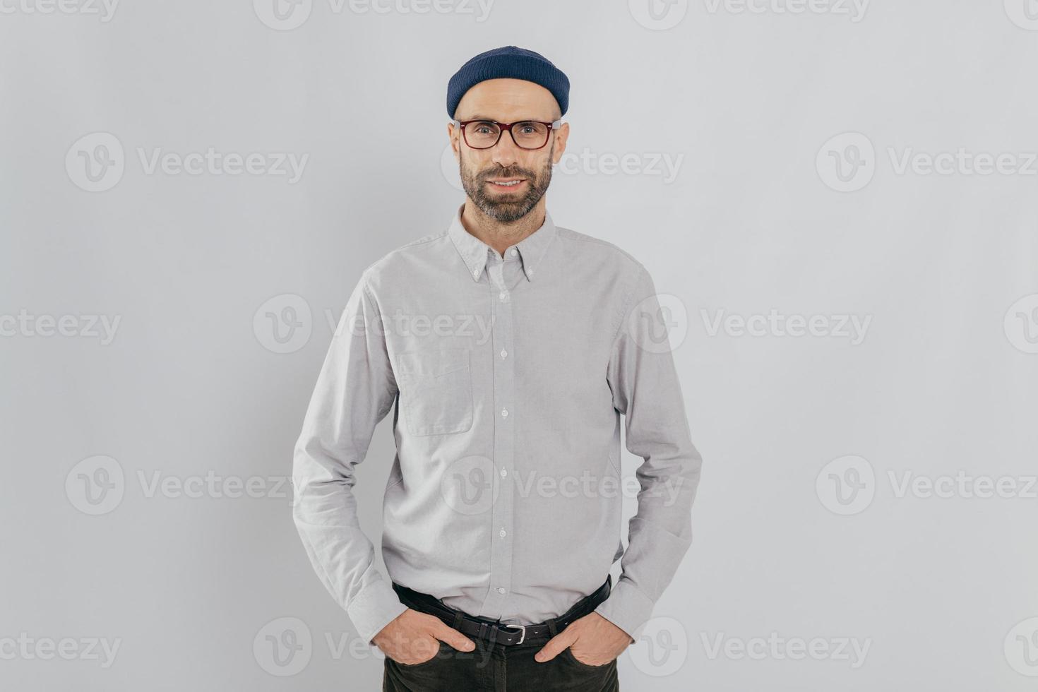 Indoor shot of satisfied Caucasian man in spectacles with bristle, keeps hands in pockets, dressed in formal stylish clothes, has gentle smile, happy to be promoted, isolated over white background. photo