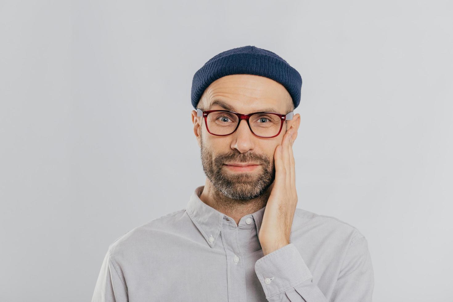 Blue eyed unshaven man raises eyebrow, keeps hand on cheek, looks happily, wears eyewear, dressed in black hat and shirt, expresses positive emotions, isolated over white wall. Facial expressions photo