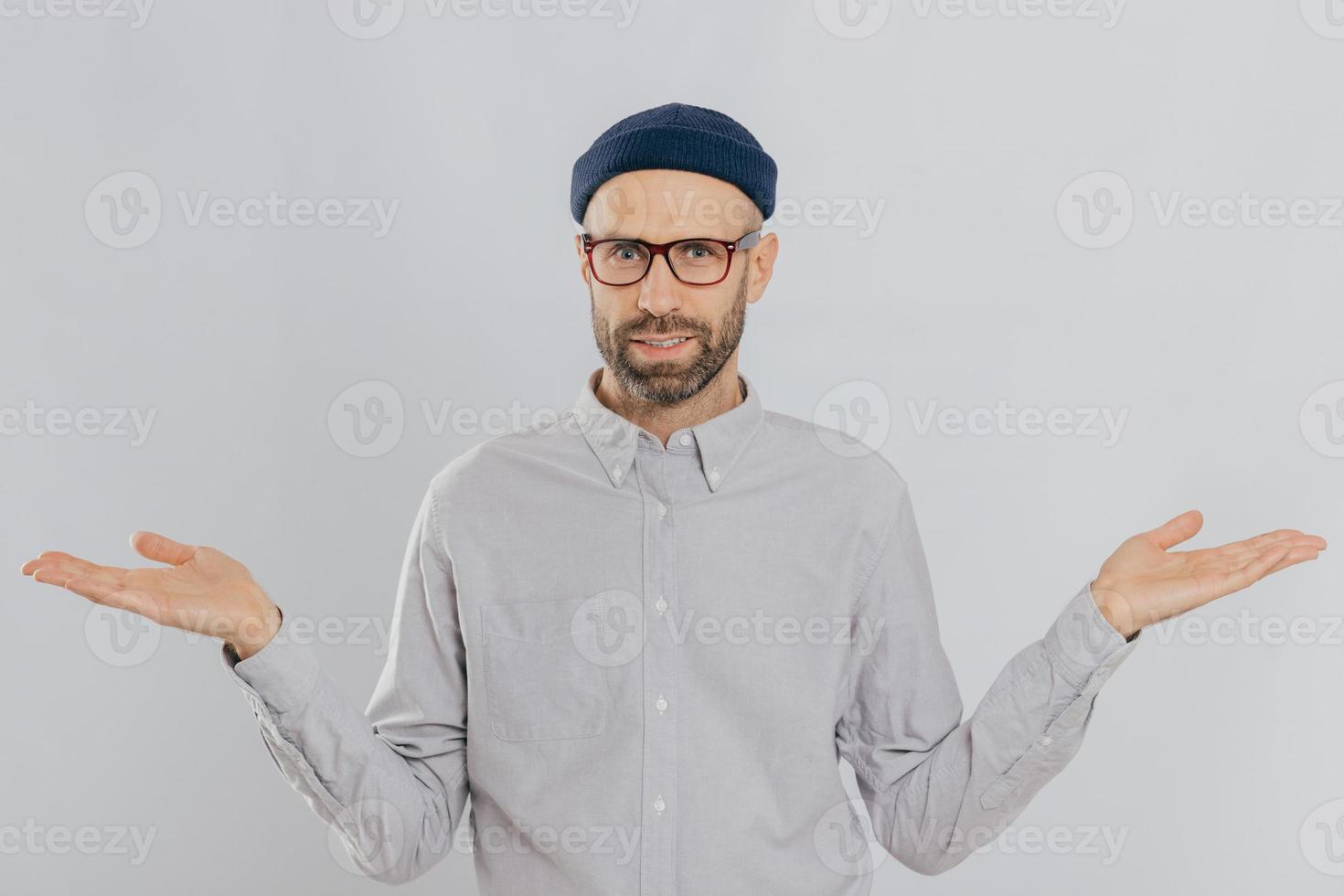 Photo of handsome man with uncertain expression, spreads hands, wears glasses and headgear, isolated over white backgrounnd. Unshaven young Caucasian male shruggs shoulders in bewilderment indoor