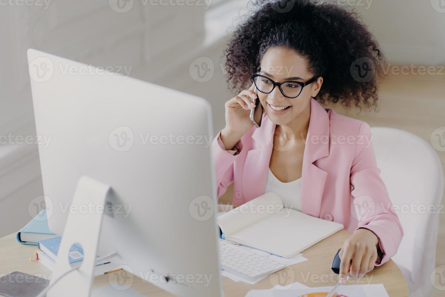 Photo of happy curly haired female wears optical glasses, pink jacket, looks attentively at computer display, sits at desktop with opened notepad, calls someone via smartphone, has positive smile