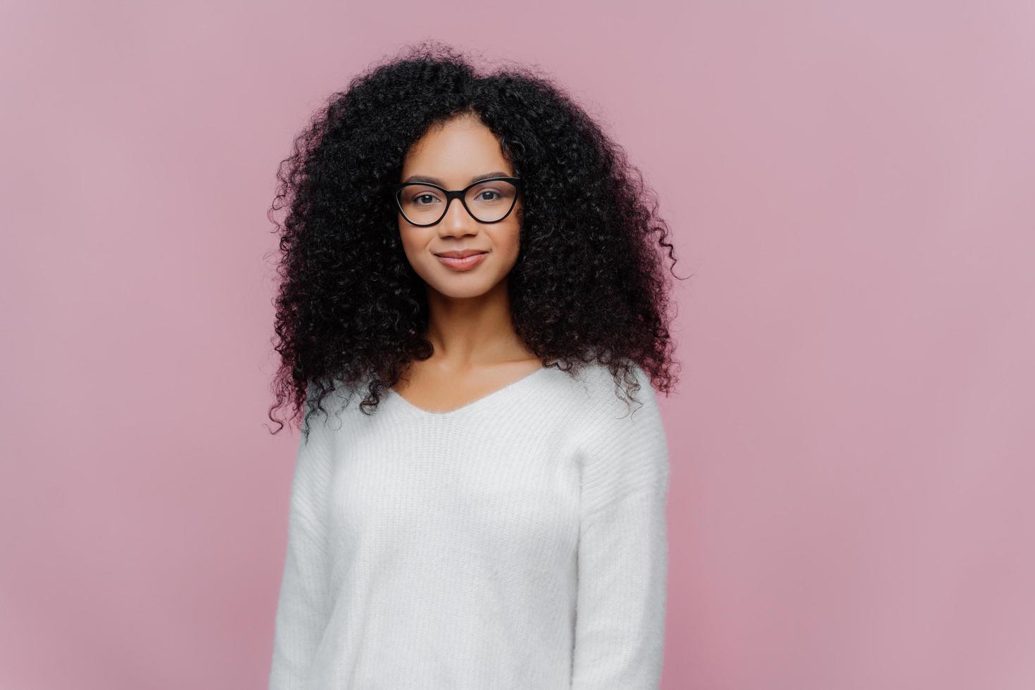 Half length shot of attractive African American woman looks through transparent glasses, white sweater, has serious calm expression, poses against violet studio wall. Facial expressions concept photo