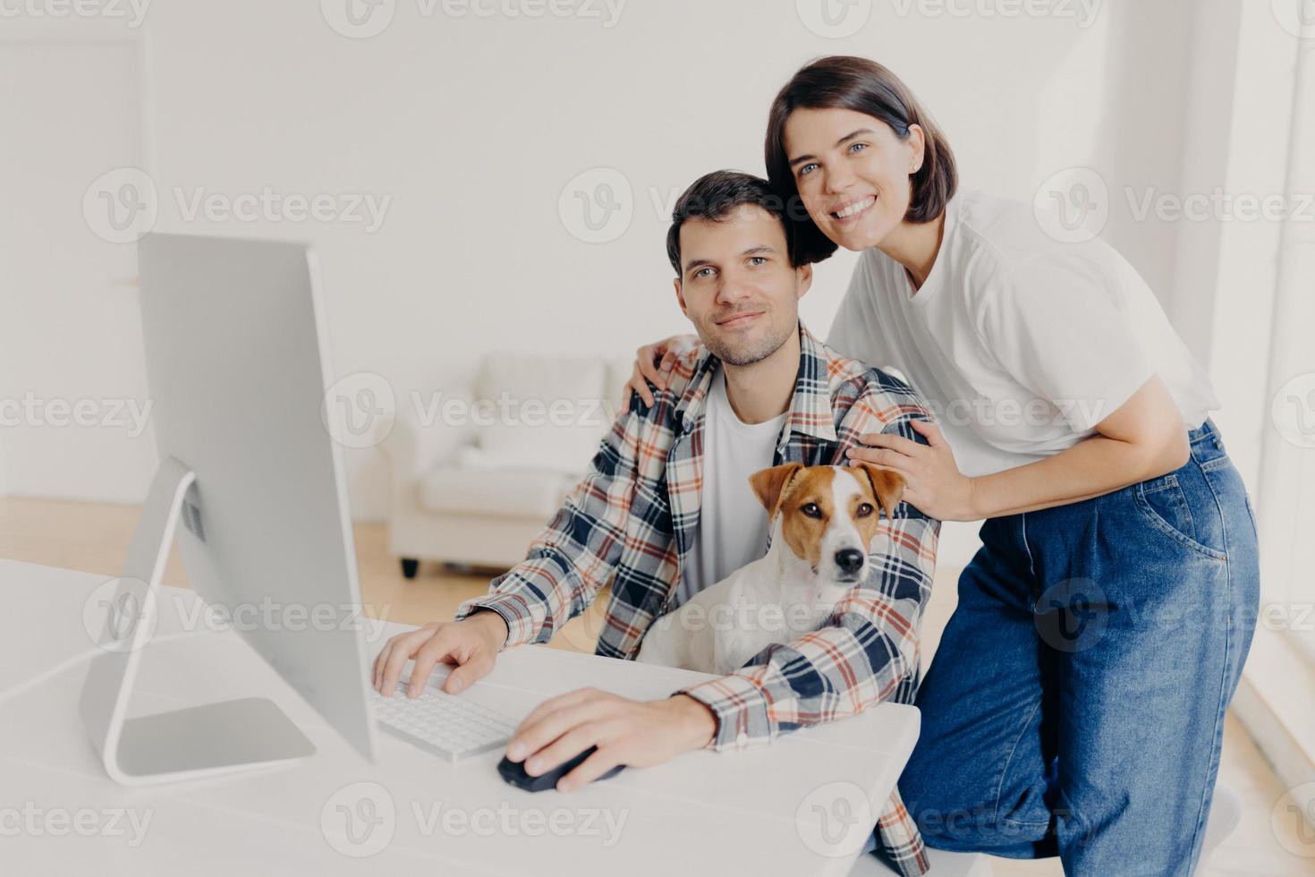 Happy husband and wife pose near computer, keyboard and surf information, work in domestic atmosphere with dog, pose in coworking space, connected to wireless internet. Lovely couple at desktop photo