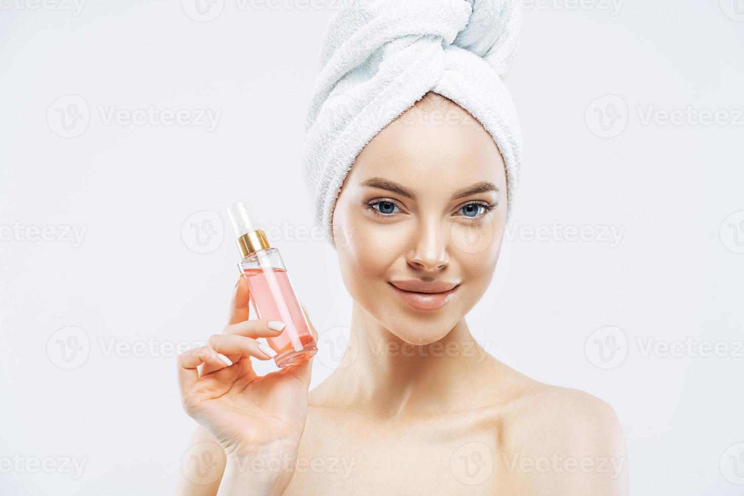 Young pleasant looking woman uses perfum, likes new smell, stands delighted indoor, applies makeup, has healthy skin wears bath towel isolated on white background has glamorous look. Pleasant smelling photo