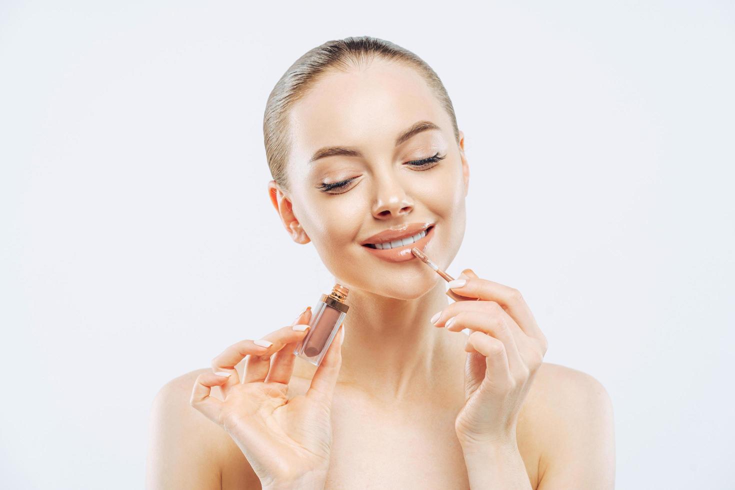 Photo of tender woman with perfect makeup, applies lip gloss, uses lip concealer brush, has dark combed hair, stands bare shoulders indoor, cares about herself, isolated on white background.