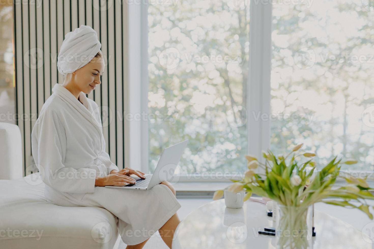 Sideways shot of attractive refreshed Caucasian woman sits at comfortbale sofa works on laptop computer wears bathrobe and wrapped towel on head poses against big window in cozy spacious room photo