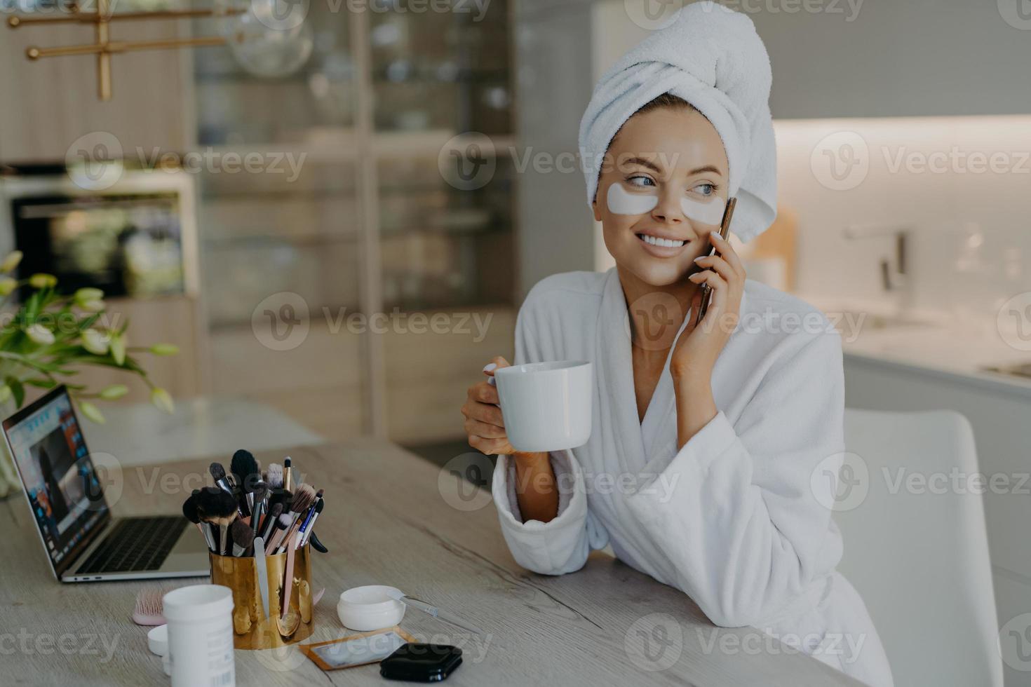 Pretty smiling woman applies hydrogel patches under eyes enjoys grooming herself after shower dressed in bathrobe drinks tea has telephone conversation sits in chair at home. Face care procedures photo