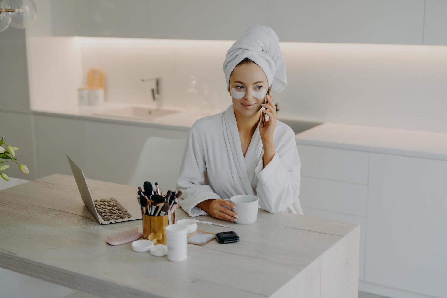 Lovely female model wears bathrobe and soft towel wrapped on head undergoes skin care procedures applies beauty patches under eyes has coffee break while talking with friend via cellular being at home photo