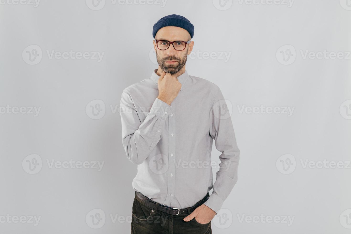 Waist up shot of serious man keeps one hand under chin, other in pocket, has stubble, wears hat, white shirt, trousers, isolated over white background, listens something attentively. Masculinity photo