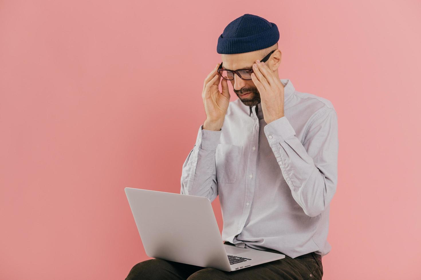Thoughtful serious student keeps hands on spectalces, has online conversation via portable laptop computer, watches webinar, wears hat and white shirt, models over pink background with free space photo