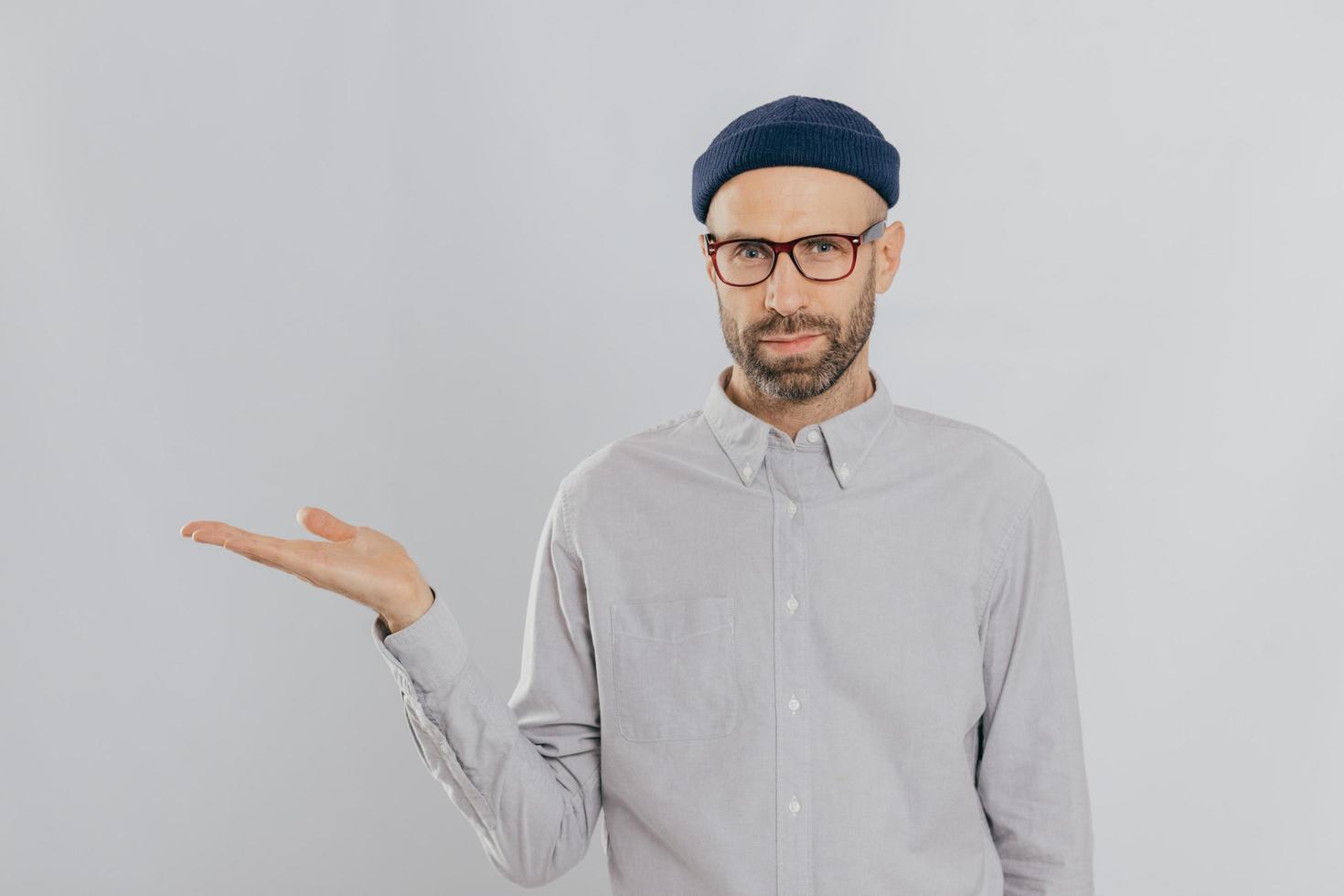 Indoor shot of unshaven Caucasian man holds copy space, demonstrates something against white wall, involved in advertising campaign. Serious male model with bristle stands alone. Isolated shot photo