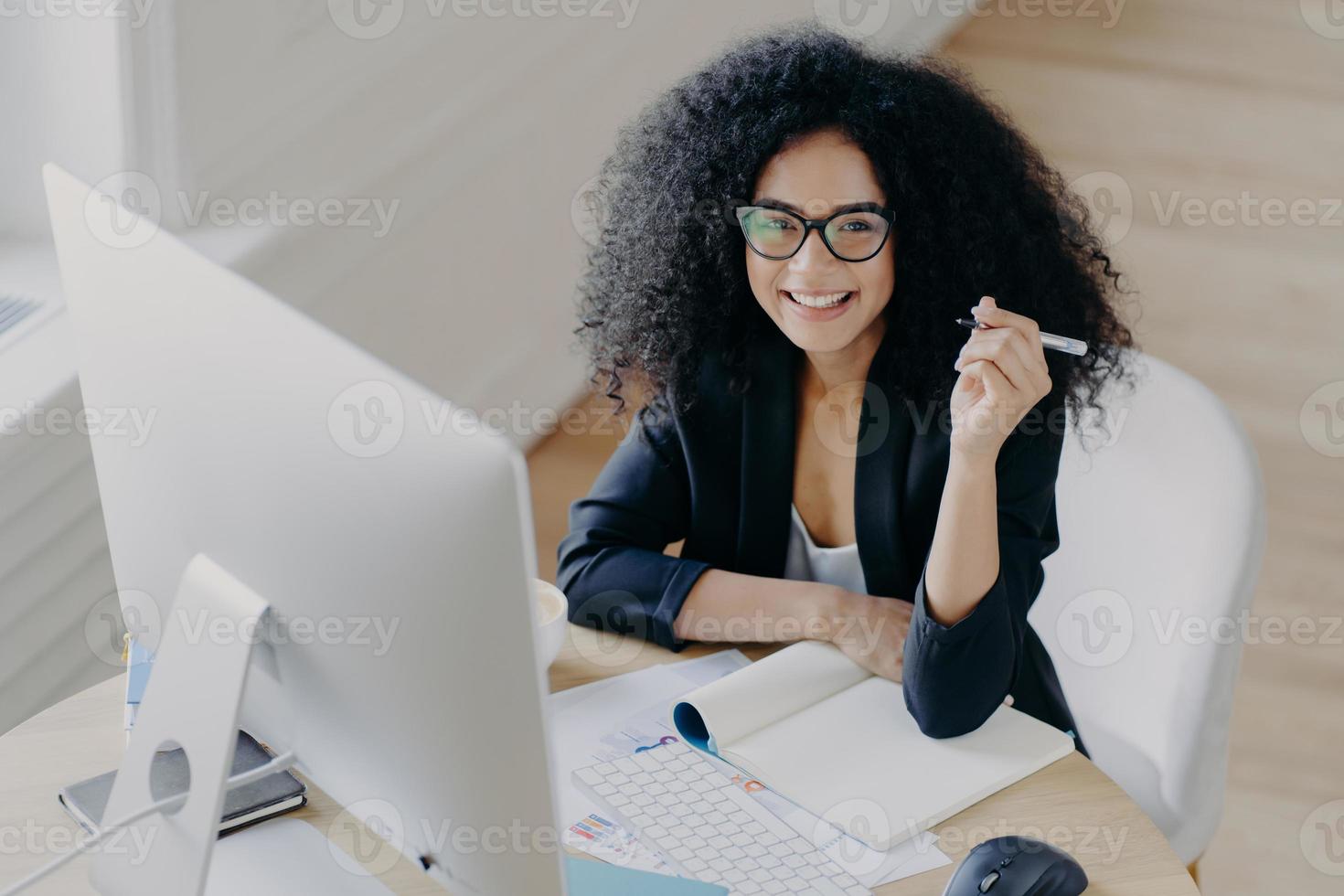 Pleased dark skinned African American woman holds pen, writes down some information, works as editor, looks gladfully at camera, holds pen, uses modern computer for searching something in internet photo