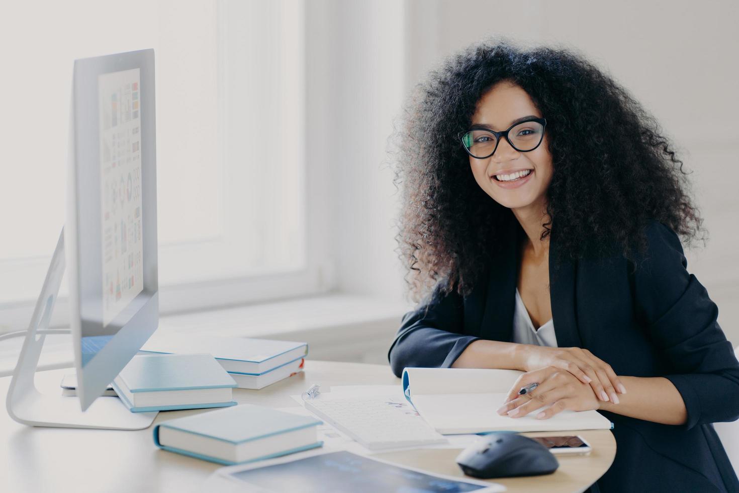 Glad curly office worker watches webinar on business topic, writes down notes with pen, smiles positively, wears spectacles and black clothes prepares report wears transparent glasses. Coworking space photo