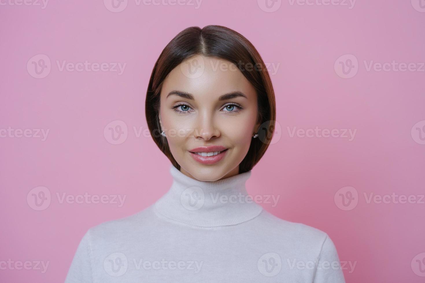 Isolated shot of charming glad young woman with pleasant smile, wears white turtleneck, looks directly at camera, isolated on pink background. Beautiful female student returns home from lectures photo