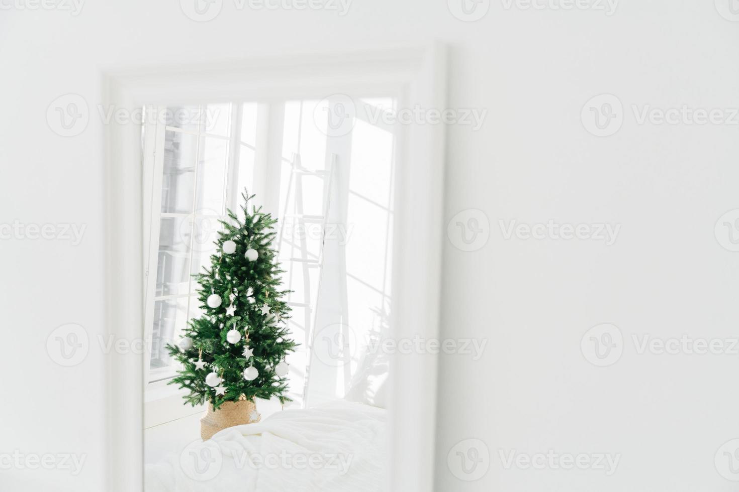 Decorated Christmas tree reflecting in mirror, white spacious room. Bedroom with New Year decoration. Modern classic style. Christmas eve at home photo