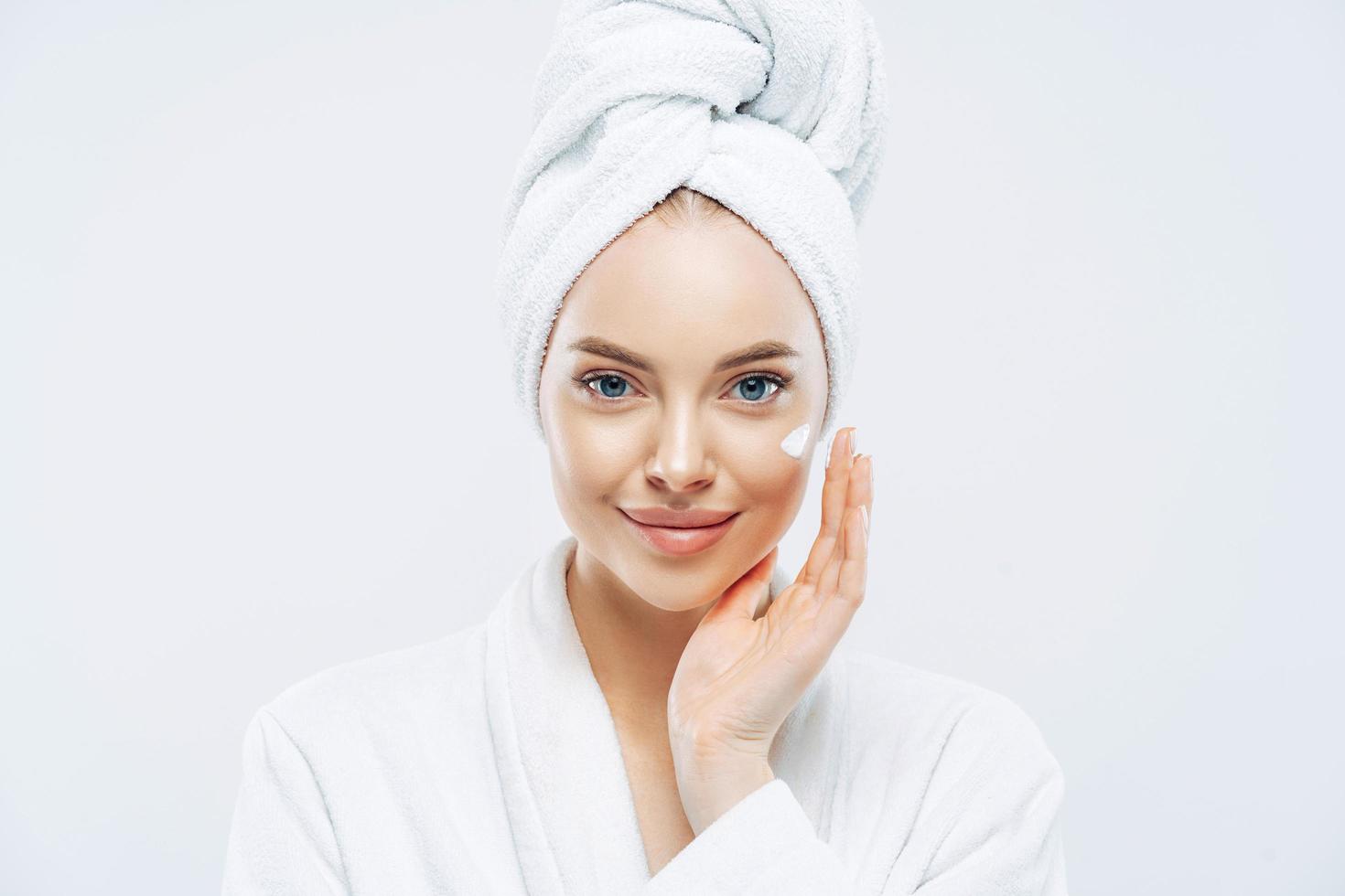 Young confident Caucasian woman applies face cream, enjoys new anti wrinkle cosmetic product, prevents sign of skin aging, wears minimal makeup, dressed in bath robe, isolated on white wall. photo