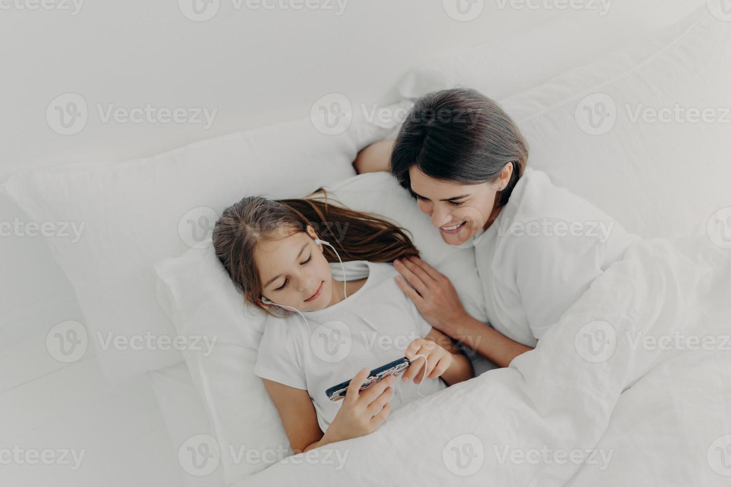 Shot of happy mother spends free time with little lovely daughter who is focused in smartphone, listens song in earphones, pose in bed with white soft bedclothes, have lazy morning during weekend photo
