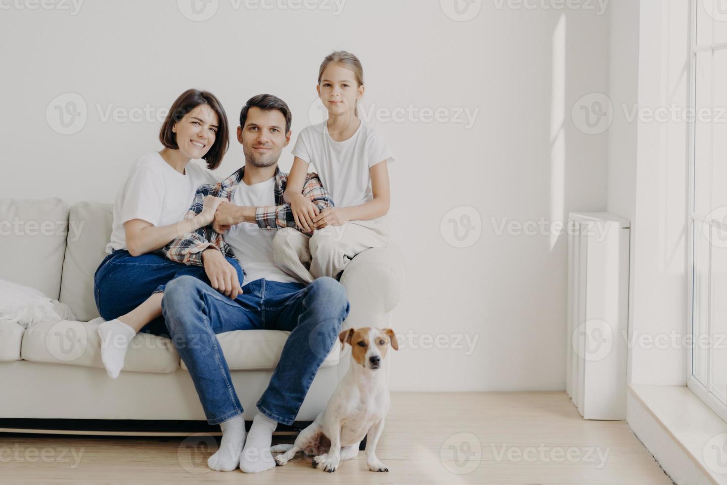 toma horizontal de una familia cariñosa posada en un sofá en una espaciosa habitación vacía con paredes blancas, su perro favorito se sienta en el suelo. copie el espacio a un lado. niña feliz contenta de estar con mamá y papá foto