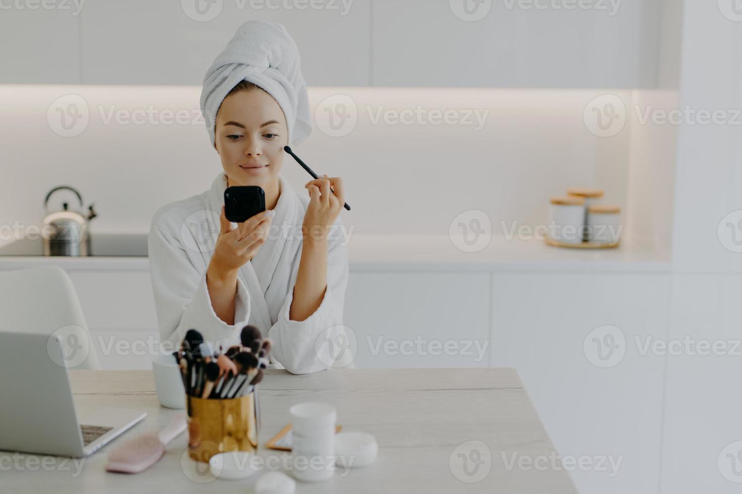 joven dama renovada aplica polvo facial con cepillo cosmético se mira en poses de espejo en la cocina en casa se sienta en el escritorio con productos de belleza usa toalla envuelta y bata blanca foto