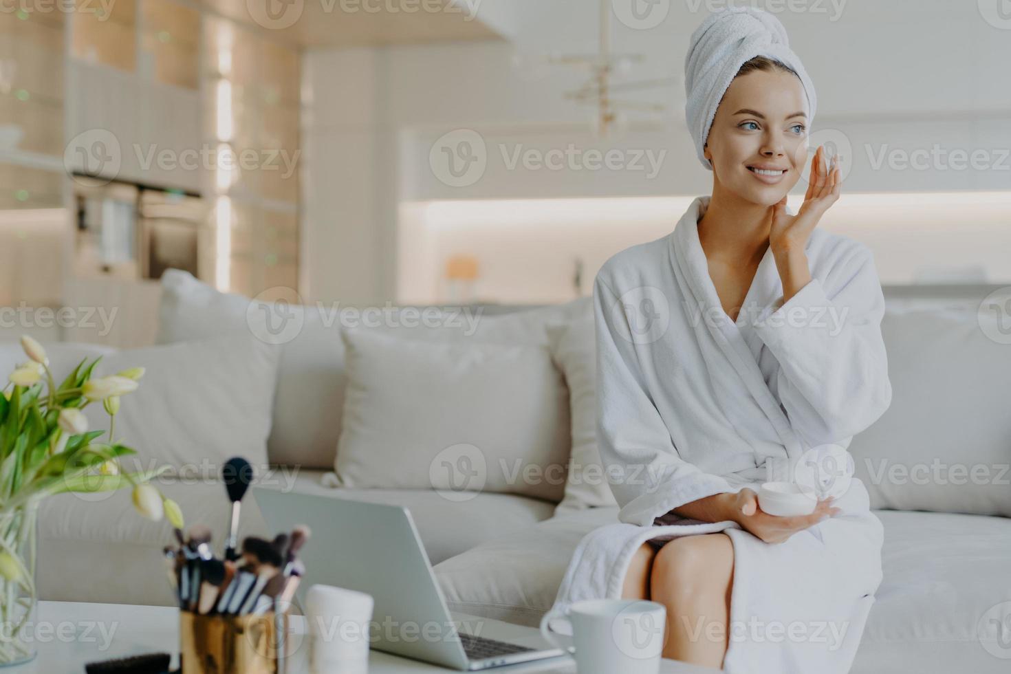 People beauty and skin care concept. Satisfied woman applies face cream enjoys facial treatments looks aside smiles gently uses cosmetic product poses at sofa in front of opened laptop computer photo